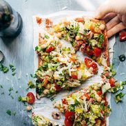 Olives, chickpeas, tomatoes, greens, herbs, and cheese on a thin crust flatbread pizza on a white plate.