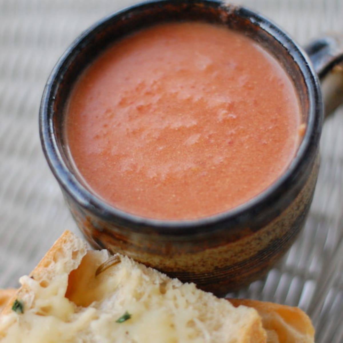 Creamy tomato balsamic soup in a mug next to a piece of bread.