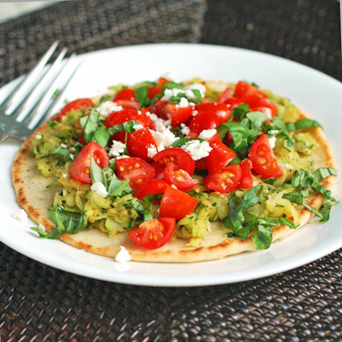 Flatbread pizza with zucchini, cherry tomatoes, and basil on a plate with a fork.